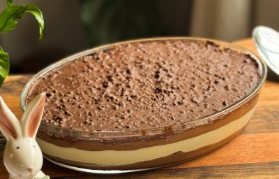 Fotografia em tons de marrom com uma travessa de vidro em formato oval ao centro. Dentro da travessa existe um pavê com camadas de chocolate meio amargo e branco, finalizadas por uma casquinha crocante de chocolate com amendoim Fotografia em tons de marrom com uma travessa de vidro em formato oval ao centro. Dentro da travessa existe um pavê com camadas de chocolate meio amargo e branco, finalizadas por uma casquinha crocante de chocolate com amendoim