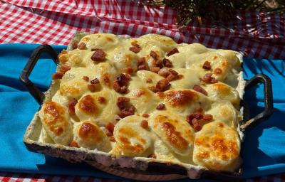 Fotografia de uma panela quadrada preta de ferro sobre um pano de mesa azul. Dentro da panela tem uma receita de batata gratinada com molho de requeijão, queijo e bacon. Fotografia de uma panela quadrada preta de ferro sobre um pano de mesa azul. Dentro da panela tem uma receita de batata gratinada com molho de requeijão, queijo e bacon.