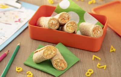 Fotografia de um recipiente laranja quadrado e fundo com enroladinhos de pão com cenoura e ovo. Dentro do recipiente tem um papel verde e branco, e ao lado tem mais dois rolinhos sobre um papel verde dobrado e uma mesa de madeira. Fotografia de um recipiente laranja quadrado e fundo com enroladinhos de pão com cenoura e ovo. Dentro do recipiente tem um papel verde e branco, e ao lado tem mais dois rolinhos sobre um papel verde dobrado e uma mesa de madeira.