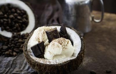 Fotografia de duas bolas de sorvete de coco com pedaços de chocolate dentro de um coco aberto, polvilhada com cacau em pó. O fruto está sobre uma mesa de madeira. Ao redor, cacau e nuts em potes pequenos na cor azul.