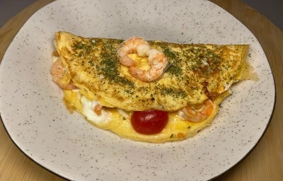 Fotografia de uma omelete dobrado ao meio com recheio cremoso de camarão e tomates cereja. Por cima da omelete tem dois camarões pequenos, e a omelete está sobre um prato raso de vidro branco.