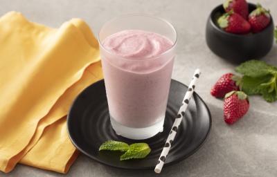 Fotografia em tons de amarelo, branco e preto de uma bancada branca com um prato preto redondo, sobre ele um copo de vidro com smoothie de morango e um canudo branco com bolinhas pretas. Ao lado um paninho laranja e morangos.