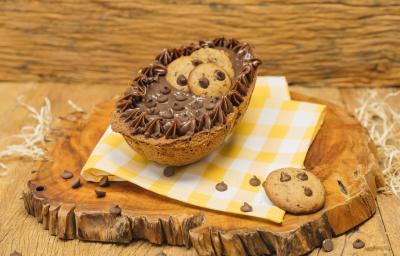 Fotografia em tons de marrom de uma bancada marrom com uma tábua de madeira com um paninho amarelo e branco, sobre ele um ovo recheado de cookie. Ao lado um cookie.