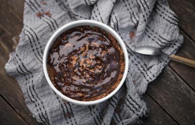 Fotografia vista de cima de um baked oats de air fryer dentro de um ramekin branco apoiado em um pano de tom cinza amassado, o qual está sobre uma madeira escura. Sobre o bolo tem uma calda de chocolate.