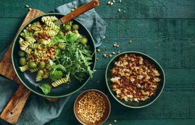 Fotografia vista de cima de uma salada verde com macarrão parafuso com iogurte, azeitona e nozes sobre uma tábua de madeira quadrada. Ao lado, dois bowls com grãos e croutons. A tábua está apoiada em um pano azul, que está sobre uma mesa em tom verde. Fotografia vista de cima de uma salada verde com macarrão parafuso com iogurte, azeitona e nozes sobre uma tábua de madeira quadrada. Ao lado, dois bowls com grãos e croutons. A tábua está apoiada em um pano azul, que está sobre uma mesa em tom verde.