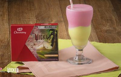 Foto aproximada de um copo alto com a receita de milkshake dividido em cores verde e rosa, servido sobre uma bancada de madeira com tecidos verde e rosa e com a caixa de KitKat Wicked ao lado esquerdo Foto aproximada de um copo alto com a receita de milkshake dividido em cores verde e rosa, servido sobre uma bancada de madeira com tecidos verde e rosa e com a caixa de KitKat Wicked ao lado esquerdo