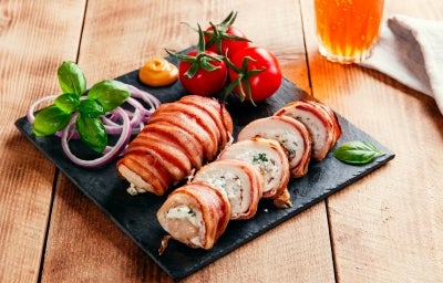 Fotografia em tons claros de dois tornedores de frango recheados enrolados no bacon, ao redor tem tomates, cebola roxa fatiada e algumas folhas, sobre um apoio quadrado preto. O apoio está sobre uma mesa de madeira.
