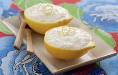 Fotografia em tons de azul, amarelo e marrom de um paninho azul estampado com um prato beje, sobre ela duas metades de um  limão com o arroz doce, uma raspa da limão. Ao lado duas colheres de madeira.