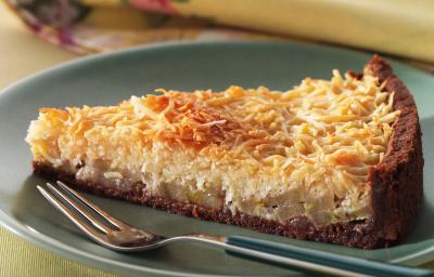 Fotografia em tons de amarelo em uma bancada de madeira, um pano amarelo com flores coloridas, um prato verde redondo raso com uma fatia da torta de coco com banana.