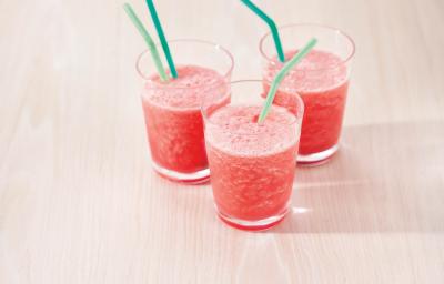 Fotografia em tons de rosa em uma bancada de madeira clara com três copos de vidro com o smoothie de melancia dentro deles e canudinhos verdes dentro dos copos. Fotografia em tons de rosa em uma bancada de madeira clara com três copos de vidro com o smoothie de melancia dentro deles e canudinhos verdes dentro dos copos.