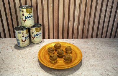 Fotografia em tons de amarelo em uma bancada bege, latas de Leite Moça ao fundo e ao centro um prato amarelo com brigadeiros.