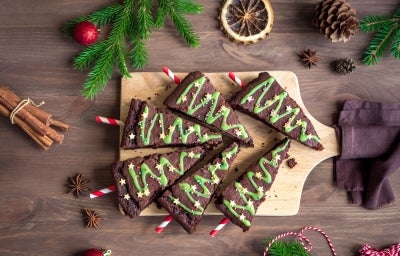 Em cima de uma mesa, tábua com brownie em forma de triângulo, decorado com creme verde e confeitos de bolinhas e estrelas.