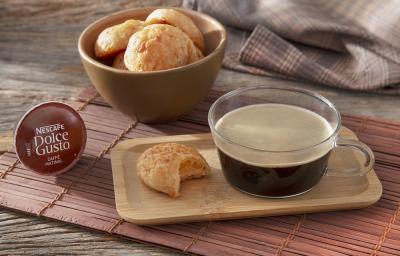 Foto da receita de pão de queijo 3 ingredientes servida em um bowl de cerâmica e à frente há uma xícara de café nescafé dolce gusto caffe matinal com um pão de queijo ao lado sobre uma tábua de madeira