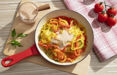 Fotografia em tons claros vista de cima de uma frigideira vermelha e branca com uma receita de moqueca e novasource. A frigideira está sobre uma tábau de madeira retangular grande ao lado de um pano brano com listras vermelhas e tomates pequenos.