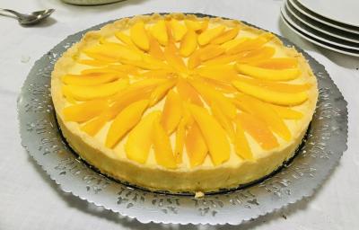 Fotografia em tons de amarelo em uma bancada de madeira de cor branca. Ao centro, uma mesa contendo a torta de manga. Ao fundo, alguns pratos brancos empilhados. Fotografia em tons de amarelo em uma bancada de madeira de cor branca. Ao centro, uma mesa contendo a torta de manga. Ao fundo, alguns pratos brancos empilhados.