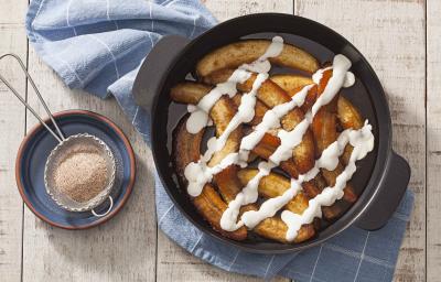 Fotografia em tons de dourado e branco, ao centro panela refratária com banana flambada e creme de leite, sobbre guardanapo azul com quadriculados brancos, ao lado potinho com uma peneirinha com açúcar e canela, sobre bancada de madeira rústica.