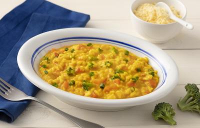 Fotografia em tons de azul em uma bancada de madeira clara com um pano azul escuro ao lado e um prato branco com detalhes em azul ao centro com o risoto de legumes com cúrcuma dentro. Ao lado, um potinho branco com queijo parmesão ralado e dois brócolis.