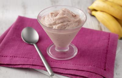 Fotografia em tons de branco e rosa de uma bancada branca com um paninho rosa, sobre ele uma colher e uma taça de vidro com o frozen. Ao fundo um cacho de banana.