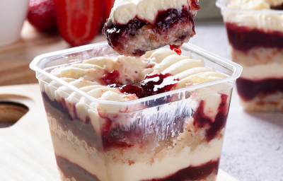 Fotografia mostra um bolo recipiente transparente mostrando camadas de um bolo de pote com creme branco e geléia de frutas vermelhas.