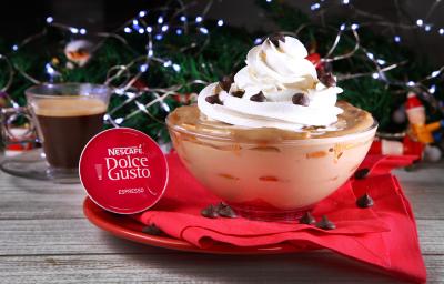 Fotografia em tons de vermelho em uma bancada de madeira, um pano vermelho em cima de um prato redondo vermelho com uma taça de vidro e o pavê de chocolate e doce de leite dentro, enfeitado com chantilly e gotas de chocolate. Ao lado, uma xícara com café.
