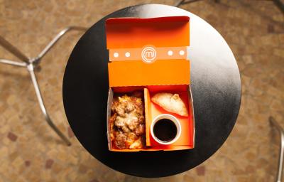Fotografia em tons de marrom com uma mesa circular preta ao centro. Em cima da mesa existe um box com uma receita de ragu de costela e um mandu frito acompanhado com um molho escuro.