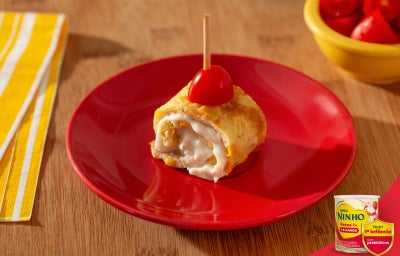 Enroladinho de omelete recheado com creme de Ninho, sobre prato vermelho, finalizado com tomate cereja e palito.