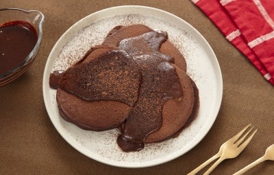 Fotografia de um prato de vidro grande e raso, branco, ao lado de alguns garfos dourados, um pano vermelho e branco e um potinho de vidro com calda de chocolate. Sobre o prato tem três panquecas de nescau com uma calda de nescau extra cacau, também.