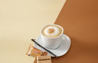 Foto da receita de cappuccino vanilla servida em uma xícara branca com um kit kat churros ao lado em fundo marrom claro e escuro