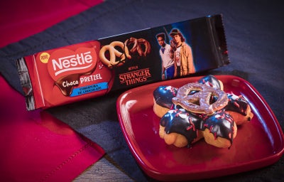 Donut em formato de flor, coberto com chocolate escuro e branco, detalhes vermelhos e um ChocoPretzel crocante no centro.