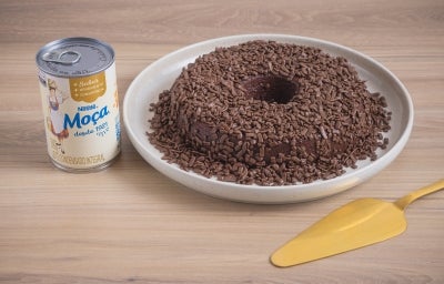 Foto da receita de Brigadeirão Moça, servido em um prato branco, decorado com muitos granulados de chocolate. O prato está entre uma lata de Leite Moça e uma espátula dourada, tudo sobre uma bancada de madeira