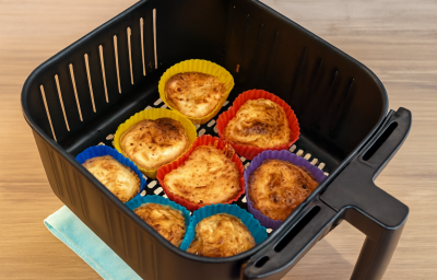 Fotografia em tons coloridos com uma bandeja de AirFryer ao centro e várias forminhas coloridas com pães de queijo dentro.
