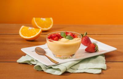 Fotografia em tons de laranja em uma bancada de madeira, um fundo laranja, um recipiente branco retangular, um pano verde claro e uma tacinha de vidro com a mousse de laranja dentro dela, com morangos para decorar. Ao fundo, uma laranja partida ao meio. Fotografia em tons de laranja em uma bancada de madeira, um fundo laranja, um recipiente branco retangular, um pano verde claro e uma tacinha de vidro com a mousse de laranja dentro dela, com morangos para decorar. Ao fundo, uma laranja partida ao meio.