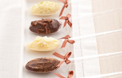 fotografia em tons de branco e marrom tirada de um recipiente retangular com 5 colheres descartáveis e todas contém uma trufa nos sabores chocolate e chocolate branco. Nas colheres um laço para decorar.