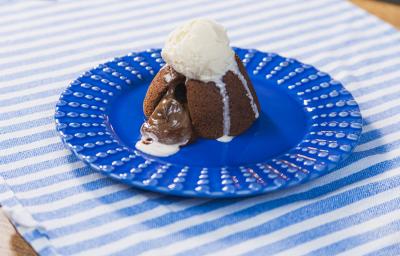 Fotografia em tons claros de um prato azul escuro raso apoiado em um pano azul claro com listras brancas. Sobre o prato tem um petit gateau aberto com a calda saindo, e uma bola de sorvete de creme escorrendo sobre a sobremesa.