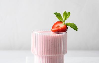 Fotografia em tons claros de de um copo de vidro com vitamina de morango sobre um apoio de mármore branca. Na borda do copo tem metade de um morango, e ao lado do copo tem mais algumas unidades dentro de um recipiente pequeno.