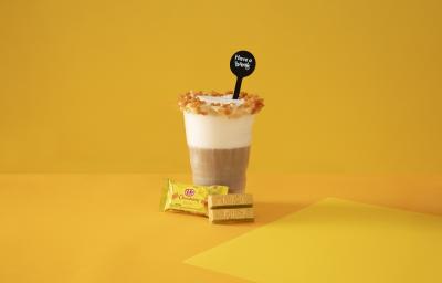 Foto em tons de amarelo da receita de latte salted caramel servida em um copo grande de vidro com um KIT KAT banana ao lado em fundo amarelo