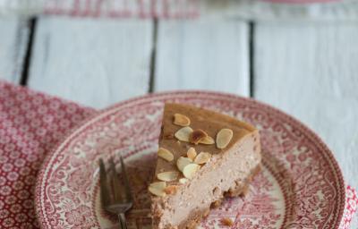 Fotografia de um prato de vidro desenhado com cor vermelho e branco. Sobre esse prato tem uma fatia de uma cheesecake de chocolate com amêndoas, ao lado de um garfo. Por cima da sobremesa tem lâminas de amêndoa. O prato está sobre um pano da mesma cor.