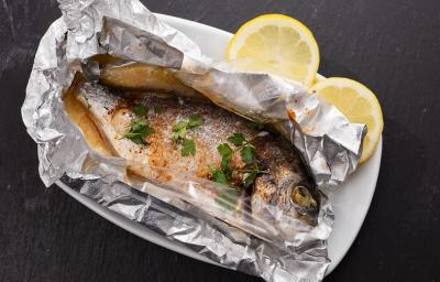 Fotografia em tons de cinza em uma bancada de madeira de cor preta. Ao centro, um prato branco contendo o peixe em um papel alumínio com duas rodelas de limão ao lado.