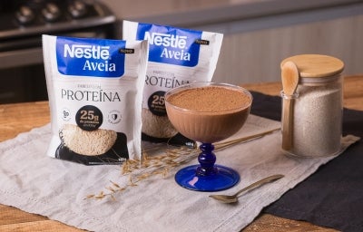 Foto da receita de mousse proteico de chocolate servida em uma taça de vidro sobre um pano neutro em cima de uma mesa de madeira com um pote de aveia ao lado com uma colher de madeira acoplada