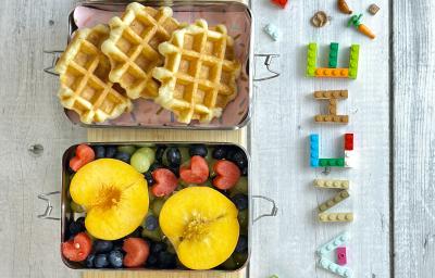 Fotografia em tons coloridos com uma lancheira com waffles e frutas dentro dela. Ao lado letras de brinquedo escrito a palara lanche em uma bancada azul em madeira. Fotografia em tons coloridos com uma lancheira com waffles e frutas dentro dela. Ao lado letras de brinquedo escrito a palara lanche em uma bancada azul em madeira.