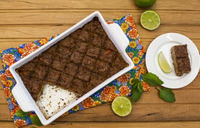 Fotografia em tons de marrom em uma mesa de madeira com um pano florido, um refratário com alças laterais brancas com o quibe de carne dentro dele. Ao lado, um pratinho branco com uma fatia do kibe e rodelas de limão. Fotografia em tons de marrom em uma mesa de madeira com um pano florido, um refratário com alças laterais brancas com o quibe de carne dentro dele. Ao lado, um pratinho branco com uma fatia do kibe e rodelas de limão.