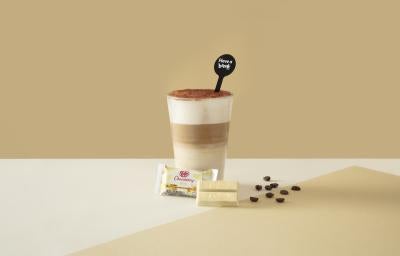 Foto da receita de Latte Tiramisú. Observa-se um fundo bege com o copo ao centro, dividido em 3 fases: embaixo, uma mais clara, a do meio, mais escura - de café, e a de cima, com creme. Por fim, chocolate polvilhado. À frente, um KITKAT de chocolate branc
