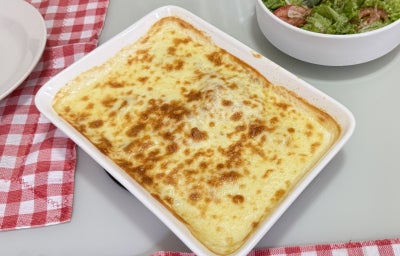 Foto da receita de tilápia gratina ao molho branco e creme de cebola, servida em uma travessa retangular branca, com um aspecto gratinado apetitoso, tudo em uma mesa com uma toalha xadrez vermelha e branca além de outro recipiente com uma salada dentro
