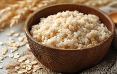 Fotografia em tons de marrom em uma bancada de madeira com um pote redondo marrom ao centro e arroz doce de aveia dentro. Ao fundo, aveia natural e um paninho marrom com uma colher de pau