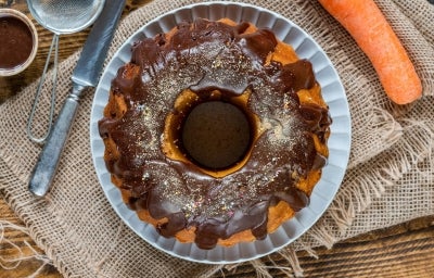 Foto vista de cima de um bolo de cenoura com cobertura de chocolate e confeitos coloridos, sobre um prato branco. O bolo está apoiado em um tecido rústico de juta, com utensílios ao redor e uma cenoura ao lado, remetendo aos ingredientes da receita. Foto vista de cima de um bolo de cenoura com cobertura de chocolate e confeitos coloridos, sobre um prato branco. O bolo está apoiado em um tecido rústico de juta, com utensílios ao redor e uma cenoura ao lado, remetendo aos ingredientes da receita.