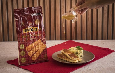 Foto da receita de Receita de Queijo Coalho Tirolez Grelhado com Cuscuz Marroquino de Ervas, Legumes Assados e Molho de Limão, servido em um prato com uma mão derramando o molho sobre ele. Ao lado do prato há uma embalagem de Queijo Coalho Tirolez.