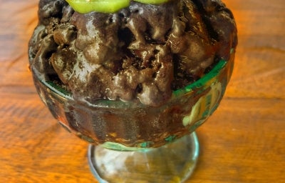 Fotografia de uma taça pequena de vidro sobre uma mesa de madeira. Dentro da taça tem uma receita de sorvete de chocolate com frutas e farinha láctea. Fotografia de uma taça pequena de vidro sobre uma mesa de madeira. Dentro da taça tem uma receita de sorvete de chocolate com frutas e farinha láctea.