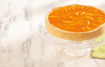 foto em tons de branco e laranja de uma bancada branca vista de cima. Contém um suporte para servir sobremesas transparente com uma torta com cobertura de tangerina por cima. foto em tons de branco e laranja de uma bancada branca vista de cima. Contém um suporte para servir sobremesas transparente com uma torta com cobertura de tangerina por cima.