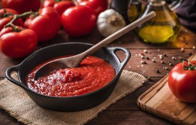 fotografia em tons de marrom e vermelho de uma bancada marrom vista de cima. Contém uma panela preta com molho de tomate e uma colher de madeira para mexer, ao lado esquerdo tomates.