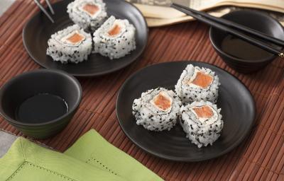 Foto em tons de marrom da receita de uramaki de salmão servida em 6 porções divididas em dois pratos pretos com um recipiente com shoyo ao lado, além de hashis pretos também. Tudo isso sobre uma esteira marrom escura com um paninho verde à frente